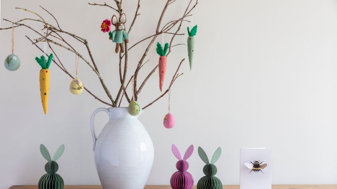 A felt rabbit, eggs and carrots hang as decorations from a vase